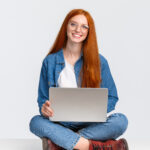 Learnteq: Attraktive, charismatische rothaarige Junge frau sitzt mit gekreuzten Beinen auf dem Boden, hält einen Laptop auf dem Schoß, arbeitet an der frischen Luft, schreibt ein neues Programm und arbeitet freiberuflich vor einem weißen Hintergrund.