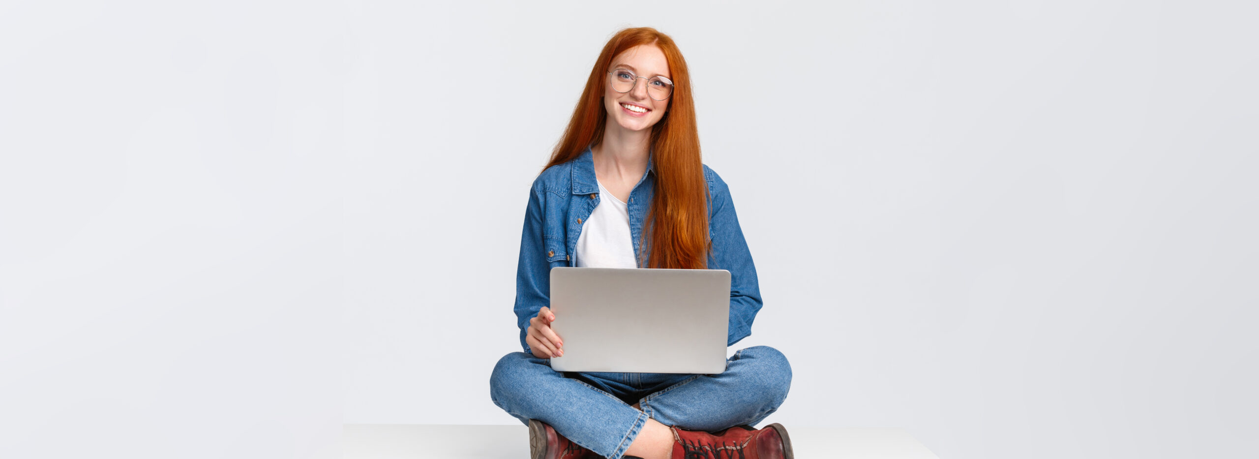 Learnteq: Attraktive, charismatische rothaarige Junge frau sitzt mit gekreuzten Beinen auf dem Boden, hält einen Laptop auf dem Schoß, arbeitet an der frischen Luft, schreibt ein neues Programm und arbeitet freiberuflich vor einem weißen Hintergrund.