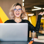 Learnteq lustige, fröhliche, begeisterte, junge, hübsche Frau, die an einem Tisch sitzt, ein schwarzes Shirt trägt, an einem Laptop in einem Coworking-Büro arbeitet und eine Brille trägt.