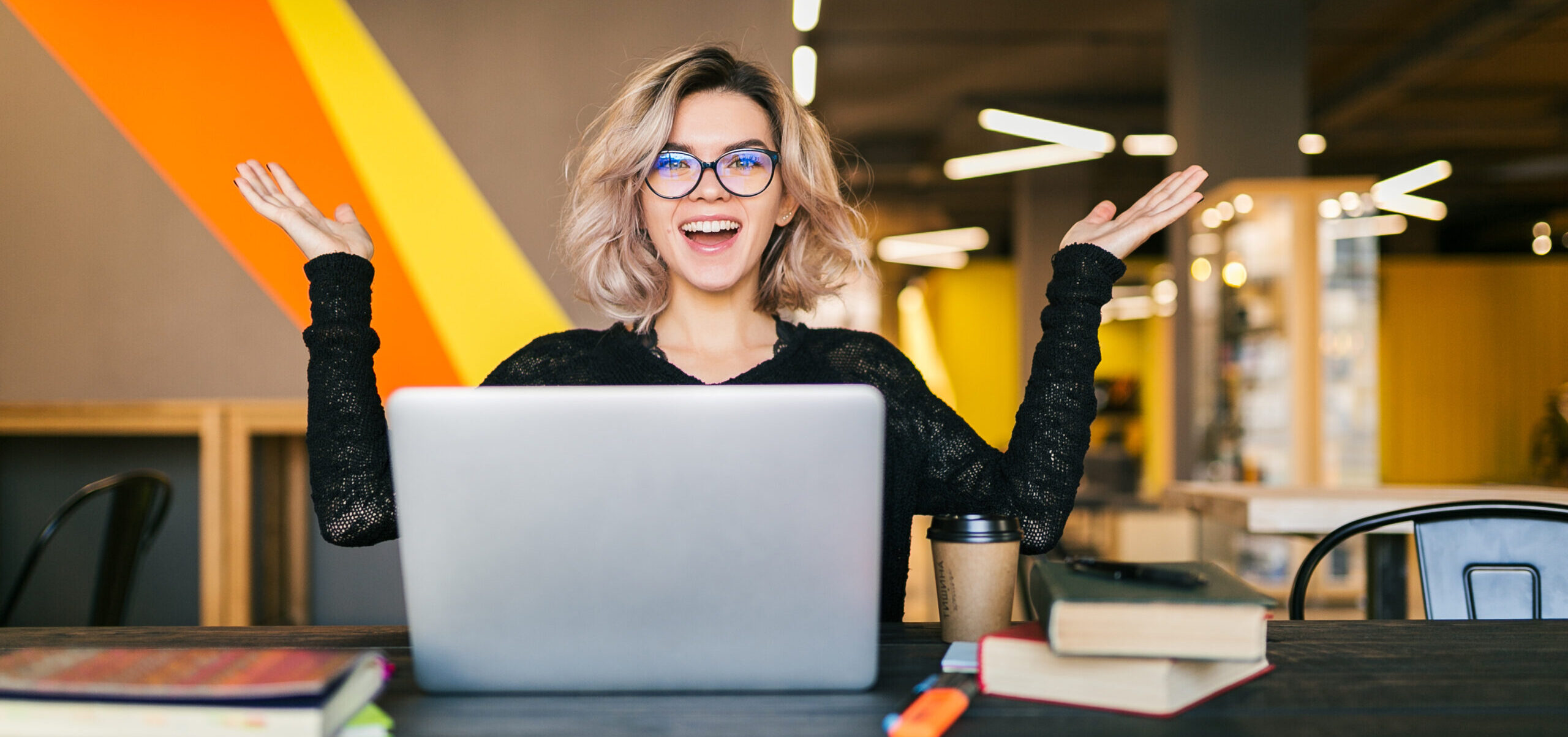 Learnteq lustige, fröhliche, begeisterte, junge, hübsche Frau, die an einem Tisch sitzt, ein schwarzes Shirt trägt, an einem Laptop in einem Coworking-Büro arbeitet und eine Brille trägt.